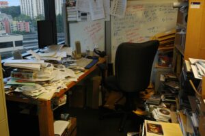Unordentlicher Schreibtisch im Homeoffice mit Papieren, Büchern und CDs – Beispiel für fehlende Ordnung am Arbeitsplatz.